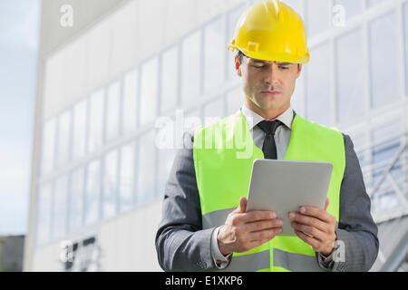 Young male engineer in reflector-vest and hardhat using digital tablet outside industry Stock Photo