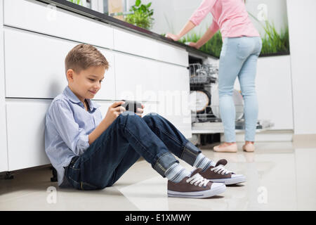 Full length of boy using hand-held video game with mother in background at kitchen Stock Photo