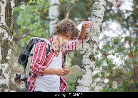 Backpacker reading map while leaning on tree trunk in forest Stock Photo