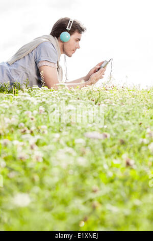 Side view of man listening to music on MP3 player using headphones while lying in park against clear sky