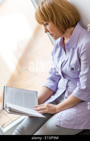 Mature woman reading book while sitting by window at home