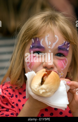 Young girl with her face painted eating a hot dog on her 6th birthday at an infant's school summer fair, Petersfield, Hampshire, UK. Stock Photo