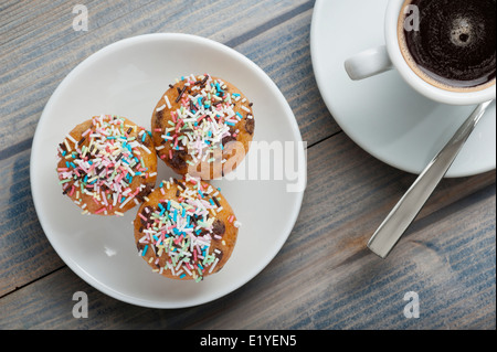 three little muffins with colorful chocolate sprinkles and cup of coffee on wooden table Stock Photo