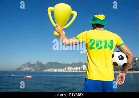 Brazilian footballer in 2014 shirt in team Brazil colors celebrating holding trophy and soccer ball Ipanema Beach Rio Stock Photo