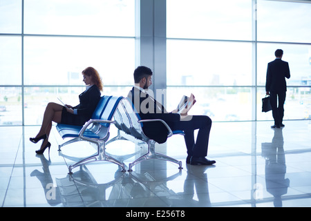 Two business partners reading at the airport on background of their colleague standing by the window Stock Photo