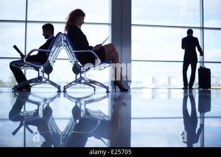 Two business partners reading at the airport on background of their colleague with suitcase by the window Stock Photo