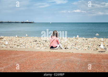 Young female sitting on her own at the beach drinking coffee, surrounded by seagulls as she enjoys the sea view Stock Photo