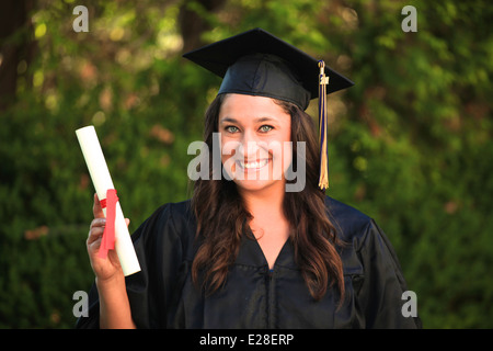 Attractive girl with blue eyes and brown hair wearing a graduation cap and gown and holding a diploma. Stock Photo