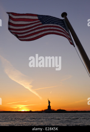 The American flag flying over the Statue of Liberty in the distance at sunset Stock Photo
