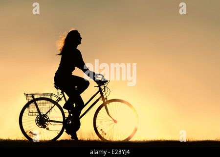 A silhouette of a happy woman outside riding a vintage bicycle, isolated in front of a sunset in the sky. Copyspace. Stock Photo