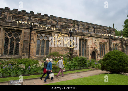 3 people walk past Holy Trinity Church, Skipton decorated with large yellow bike & bunting flags celebrating Tour de France - Yorkshire. England, UK. Stock Photo