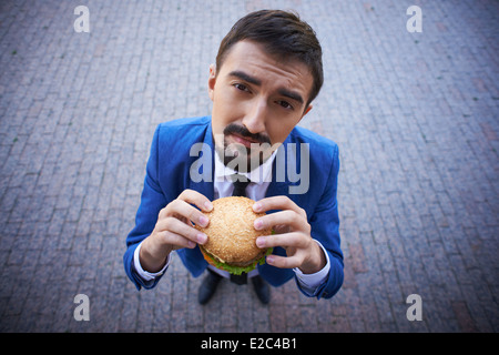 Image of hungry businessman with sandwich looking at camera outside Stock Photo