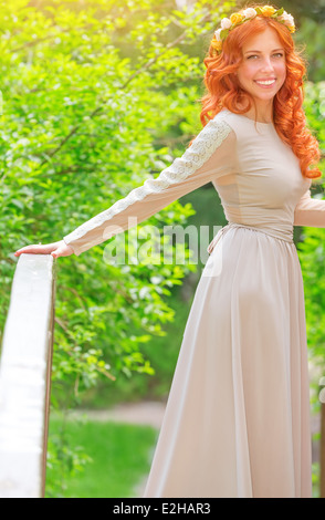 Beautiful happy bride wearing long beige dress and flower wreath in red hair, standing on little bridge in fresh summer park Stock Photo