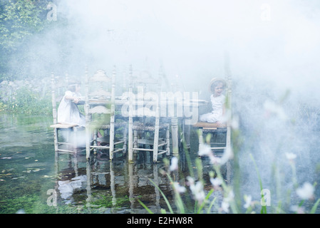 Children sitting at dining table floating on pond, surrounded by smoke Stock Photo