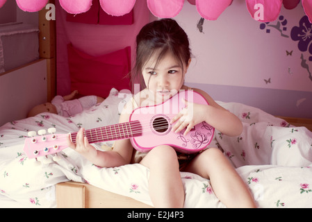 Little girl sitting on bed, playing toy guitar