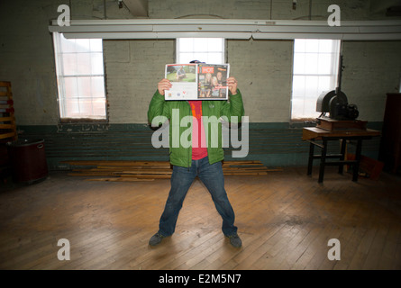 A 40-year-old man wearing a green jacket, red shirt, and jeans reads ? while standing in a room in a corset factory. Stock Photo