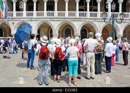 Group of tourists on Venice tour standing & listening to guide (hand raised) outside Doges Palace museum on very hot sunny day thus the sun hats Stock Photo