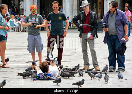 People chatting to woman laying down on paving slabs in St Mark's Square Venice feeding the pigeons from her hands Stock Photo