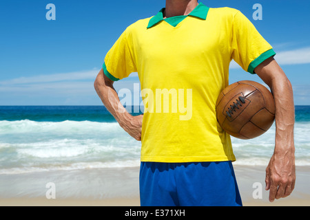 Brazilian football player in team Brazil colors holding old vintage brown soccer ball on tropical beach Stock Photo