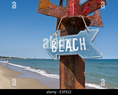 Wooden beach sign hanging on rustic metal post with couple in background strolling at waters edge Sandbanks Dorset UK Stock Photo