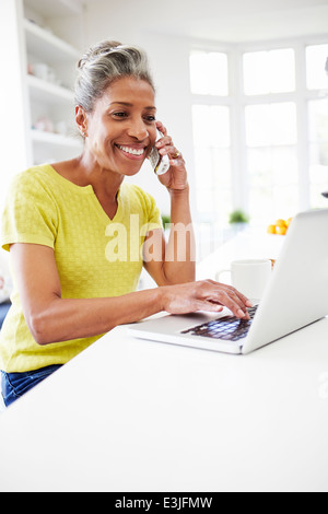 Woman Using Laptop And Talking On Phone In Kitchen At Home Stock Photo