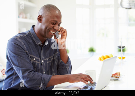 Man Using Laptop And Talking On Phone In Kitchen At Home Stock Photo