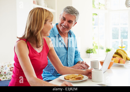 Couple Using Laptop Whilst Having Breakfast In Kitchen Stock Photo