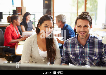 Two Colleagues Working At Desk With Meeting In Background Stock Photo