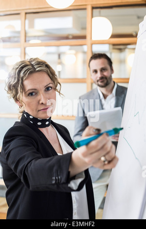 Portrait of mature businesswoman writing on flip chart with male colleague in background at office Stock Photo
