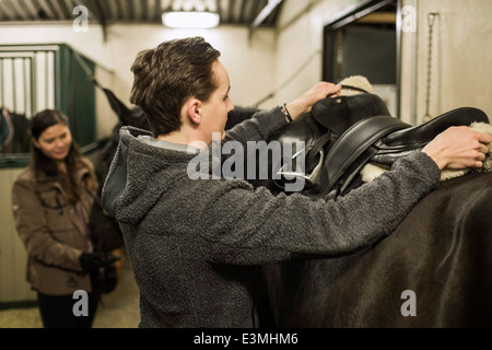 Young man preparing horse in stable with woman in background Stock Photo