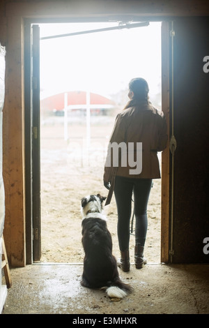 Full length rear view of young woman with dog leaning in doorway of horse stable Stock Photo