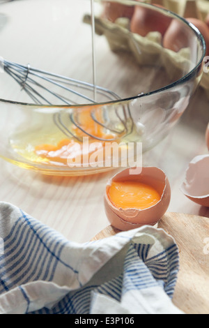Cracked egg atop wooden kitchen counter. Close up of raw egg with glass bowl, whisk and egg carton in the background. Stock Photo