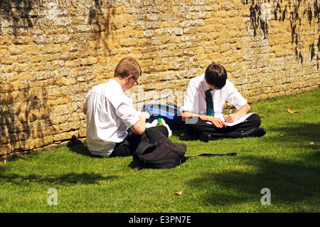 Two teenage schoolboys sitting on the grass by a Cotswold stone wall doing their schoolwork, Bourton on the Water, England, UK. Stock Photo