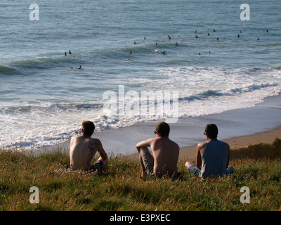 Three young men sat on the cliff top looking out to sea, Widemouth Bay, Bude, Cornwall, UK Stock Photo