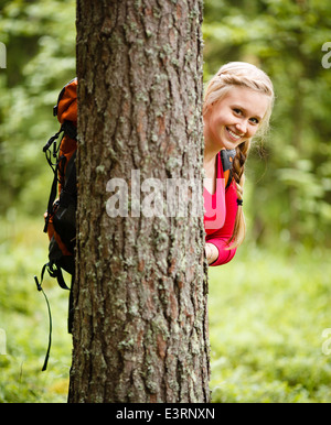 Young blond woman hiking and hiding behind a tree in the forest. Stock Photo