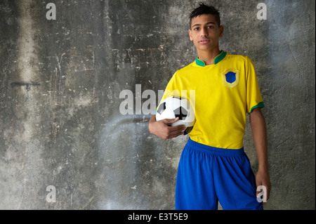 Young Brazilian footballer in team Brazil colors kit holding soccer ball against concrete favela wall Stock Photo