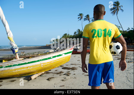 Brazilian football player in 2014 shirt colors holding soccer ball in front of fishing boat on Nordeste Brazil beach Stock Photo