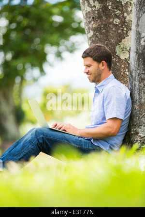 Young man working outside on laptop in park under a tree Stock Photo