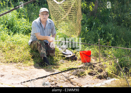 Old man sitting on riverbank fishing at daytime Stock Photo