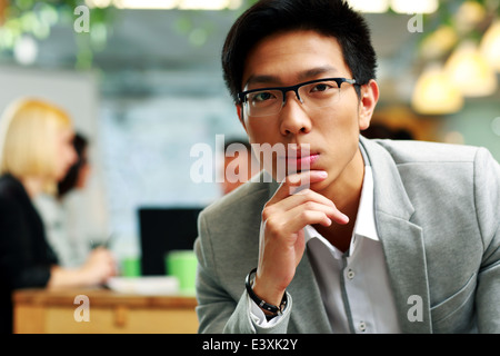 Portrait of a thoughtful asian man sitting in office, with his colleagues in background Stock Photo