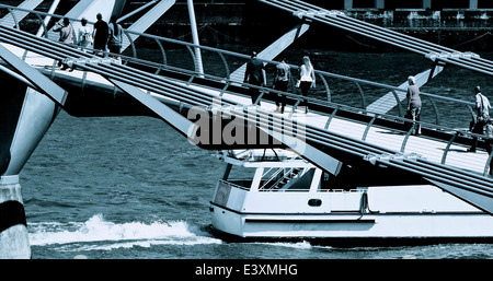 Tourists and commuters on the Millennium Bridge as boat speeds past travelling eastwards on the river Thames London England Stock Photo
