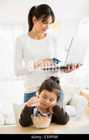 Hispanic mother shopping online while daughter eats cereal