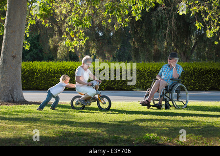 Three year old boy pushing grandmother on cycle with grandfather watching from wheelchair Stock Photo