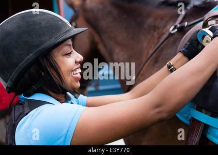 Close up of young woman putting saddle on horse Stock Photo