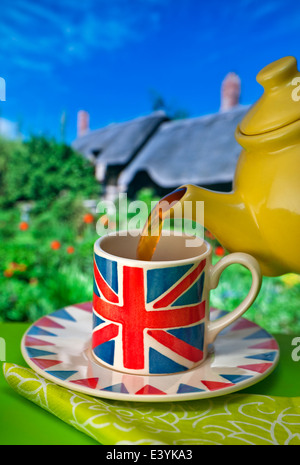 English Tea Staycation Union Jack themed cup, yellow china teapot pouring tea with typical thatched cottage and garden in background UK Stock Photo