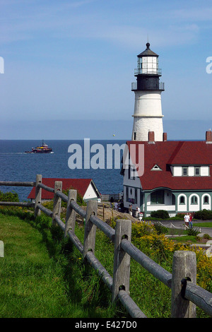 Portland Head lighthouse guides tug boat in Maine. It is located in Fort Williams Park. Wooden fence leads to beacon. Stock Photo