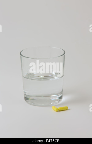 Two yellow tablets next to a small, clear glass of water with a plain white background Stock Photo