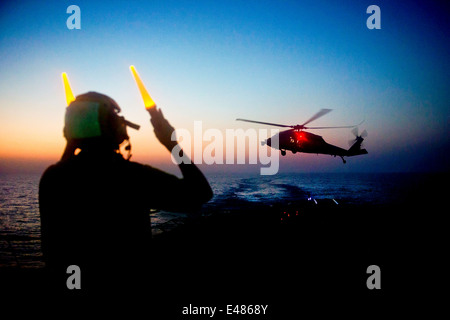 US Navy sailor signals to pilots in an MH-60S Sea Hawk helicopter to land on the flight deck of the guided-missile destroyer USS Arleigh Burke during night time replenishment June 20, 2014 in the Arabian Sea. Stock Photo