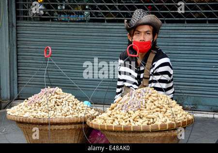 BANGKOK, THAILAND: Young man wearing a cowboy hat and a red mask sitting on the sidewalk at Thanon Ratchaburi selling peanuts Stock Photo