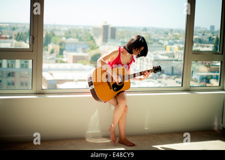 Young woman sitting on window sill and playing guitar Stock Photo